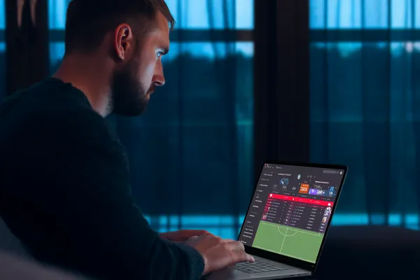 একজন পুরুষ তার ল্যাপটপে মনোযোগ দিয়ে কাজ করছেন, ব্যাকগ্রাউন্ডে লাইভ স্ট্যাটস — যা A71 মোবাইল অ্যাপের স্পোর্টস ও গেমিং ফিচারগুলোকে তুলে ধরে।